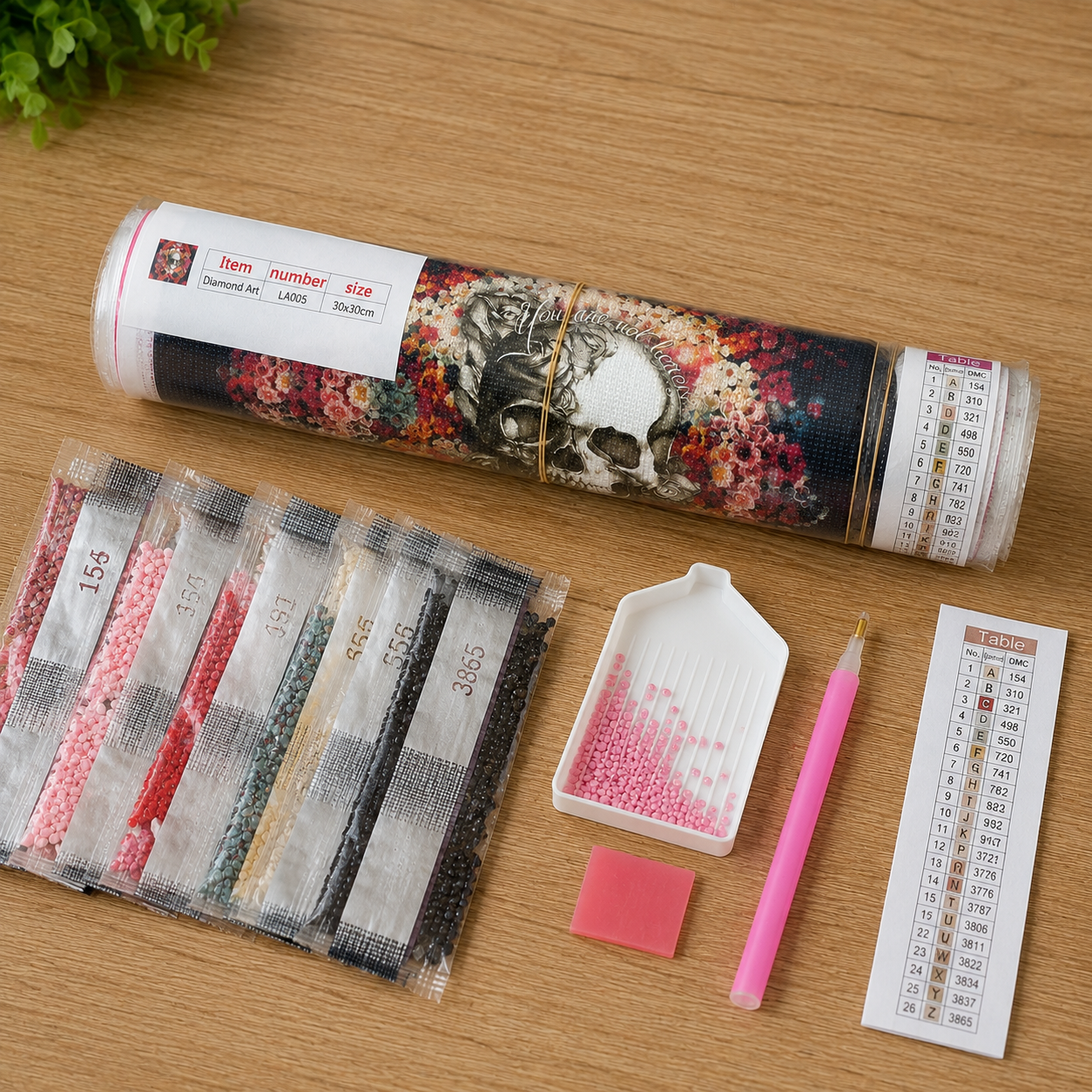 Floral Mandala Skull Diamond Painting Kit – flat lay on warm wood surface showing floral mandala skull canvas rolled in a clear tube labeled Diamond Art LA005 30x30cm, 8 resealable diamond drill bags in pink, red, black, teal, cream, and dark tones, white grooved sorting tray with pink drills, pink applicator pen, pink wax pad, and DMC color chart reference card