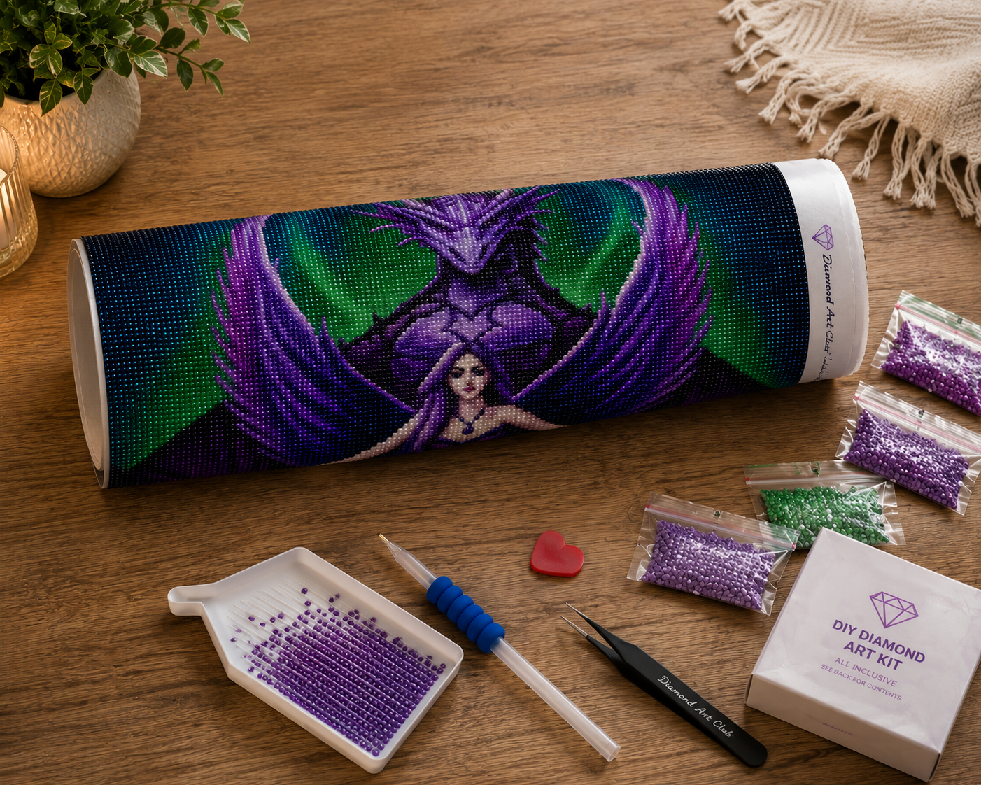 Purple Dragon Guardian & Angel Diamond Painting Kit – flat lay on warm wood surface showing purple dragon angel canvas rolled in a Diamond Art Club tube, resealable diamond drill bags in purple, violet, green, and lavender tones, white grooved sorting tray with purple drills, blue applicator pen, black Diamond Art Club tweezers, red heart wax pad, and white DIY Diamond Art Kit box