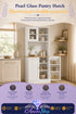 White pantry hutch with framed glass doors, styled in a warm beige kitchen corner with dishware, jars, and greenery.
