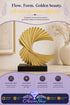 A gold metallic sculptural wave figurine displayed on a black rectangular base, set against a warm beige interior background with a vase and framed art. The image includes promotional text and a purple footer banner with the brand name "Azurra Shop".